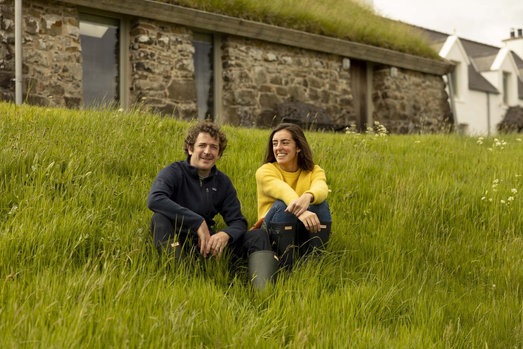 James Mitchell and Carolina Larrazábal sitting on the grass in front of the Blackhouse at Mint Croft, Isle of Skye