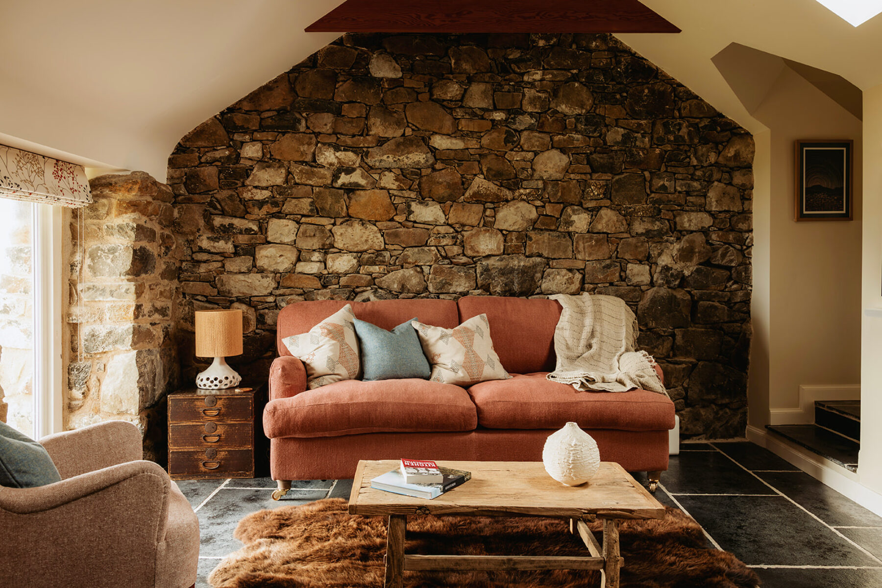 Inside the historic blackhouse holiday cottage, a traditional hebridean building in the Isle of Skye at Mint Croft. An exposed stone wall and flagstone floor with comfortable, upholstered sofa and armchair in warm tones. On the floor a sheepskin rug.