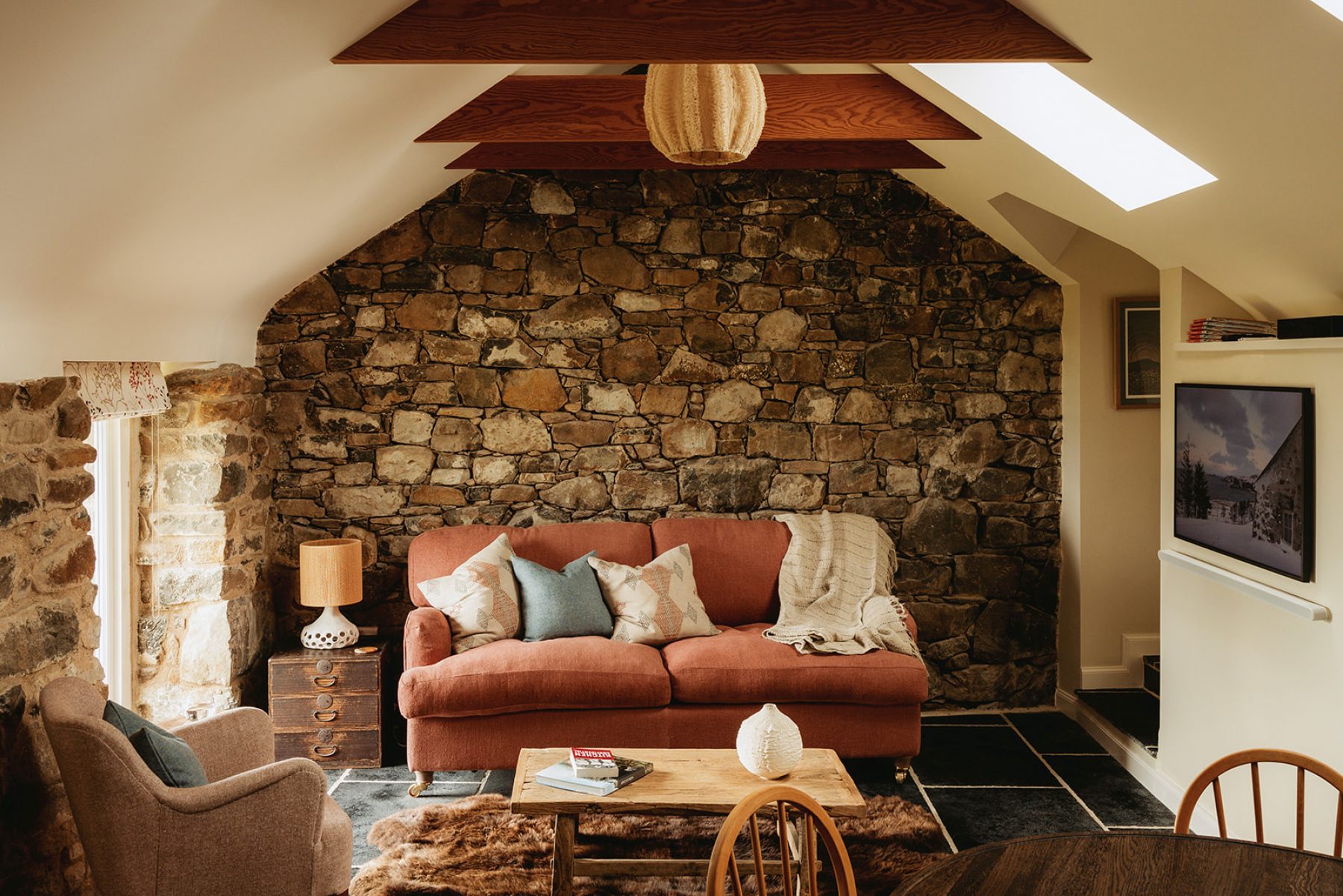 The sitting room at the Blackhouse cottage, Mint Croft, Isle of Skye. Exposed rafters and stone wall in this traditional croft building with luxury self-catering accommodation. Comfortable sofa, chair and soft furnishings in soft oranges and blues.