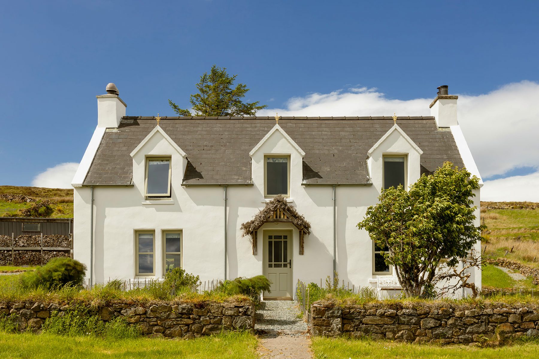 Traditional Scottish Highland croft house cottage at Mint Croft, Skye. A stone whitewashed house with three dormer windows upstairs, a pitched slate roof and a heather-roofed porch above the front door. Set behind a herb garden with low dry stone wall. A bright early summer day with blue sky