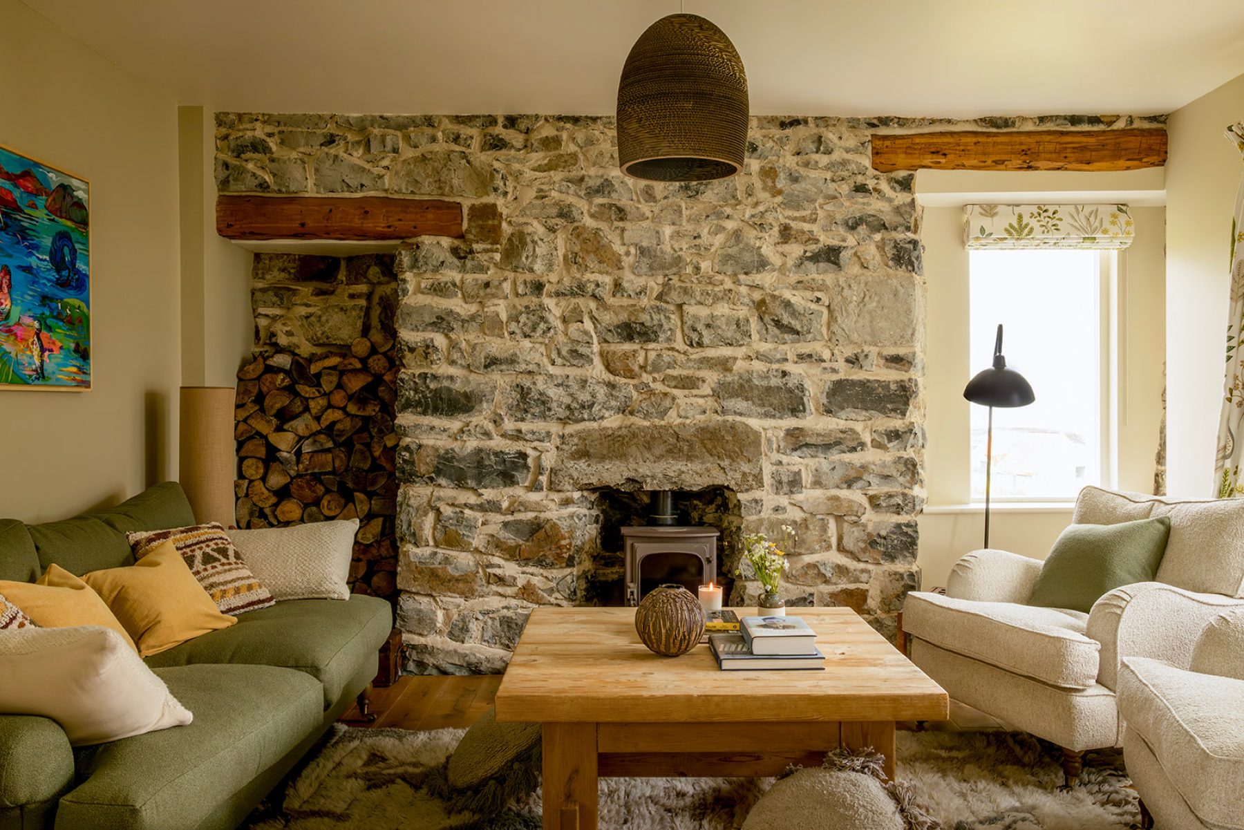 Croft House interior, sitting room at Mint Croft, Skye, Scotland. An exposed stone wall with wood burner and wood store, comfortable upholstered furniture, wooden coffee table.