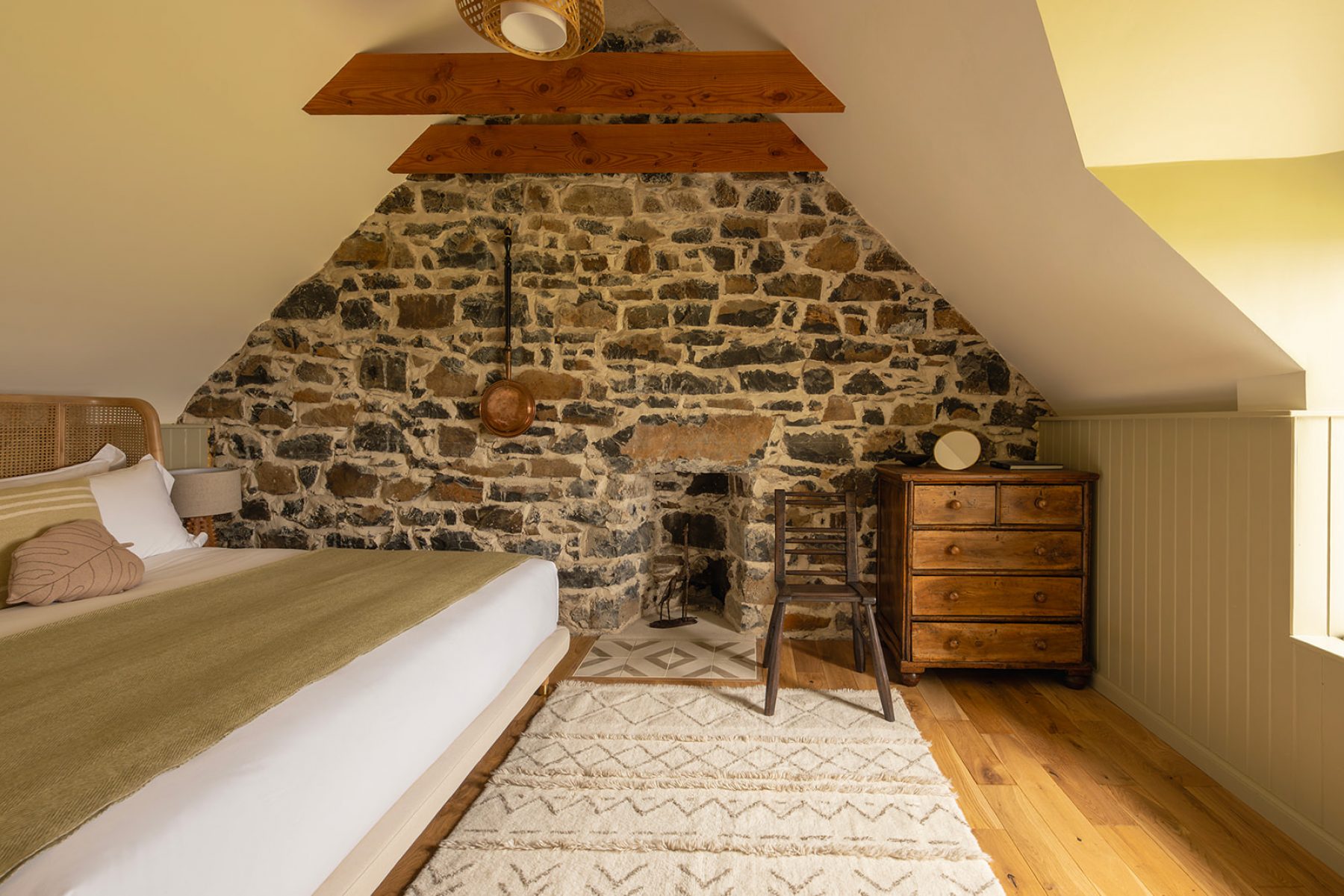 Croft House bedroom with exposed stone wall at the gable end. A large bed with curved wicker headboard, crisp linen and a green blanket. Antique chest of drawers and a contemporary Caithness chair by Lawrence Veitch