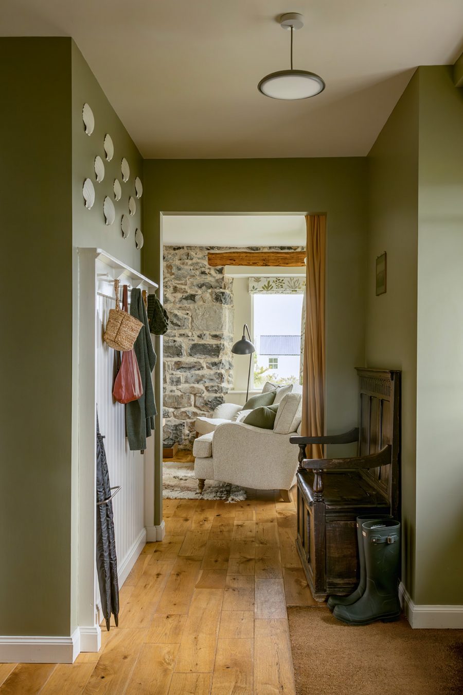 Croft House entrance hall at Geary, Skye, Scotland. Soft sage green walls with an antique wooden settle, tongue and groove with pegs for coats, scallop shelves on wall. Leading through to sitting room with exposed stone wall.