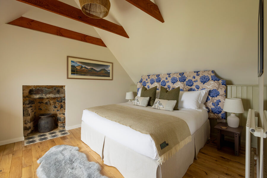 Croft House bedroom at Mint Croft, Isle of Skye. A large bed with upholstered headboard in floral fabric, crisp bedlinen. An open hearth with expose stone. Exposed wooden roof trusses