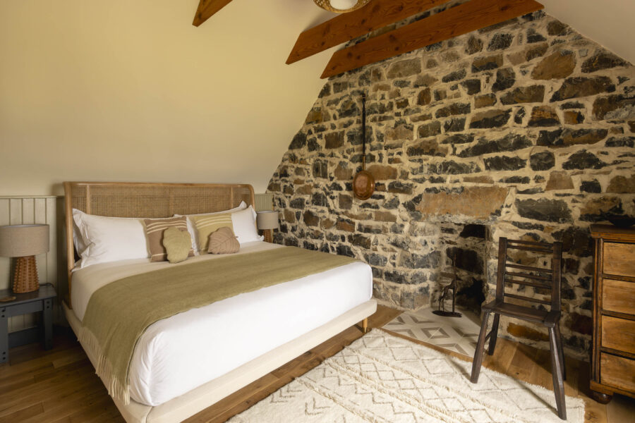 Bedroom with exposed stone walls in the traditional Highland Croft House cottage at Mint Croft Skye, Scotland. A large double bed with wicker headboard and a contemporary Caithness chair by Lawrence Veitch.