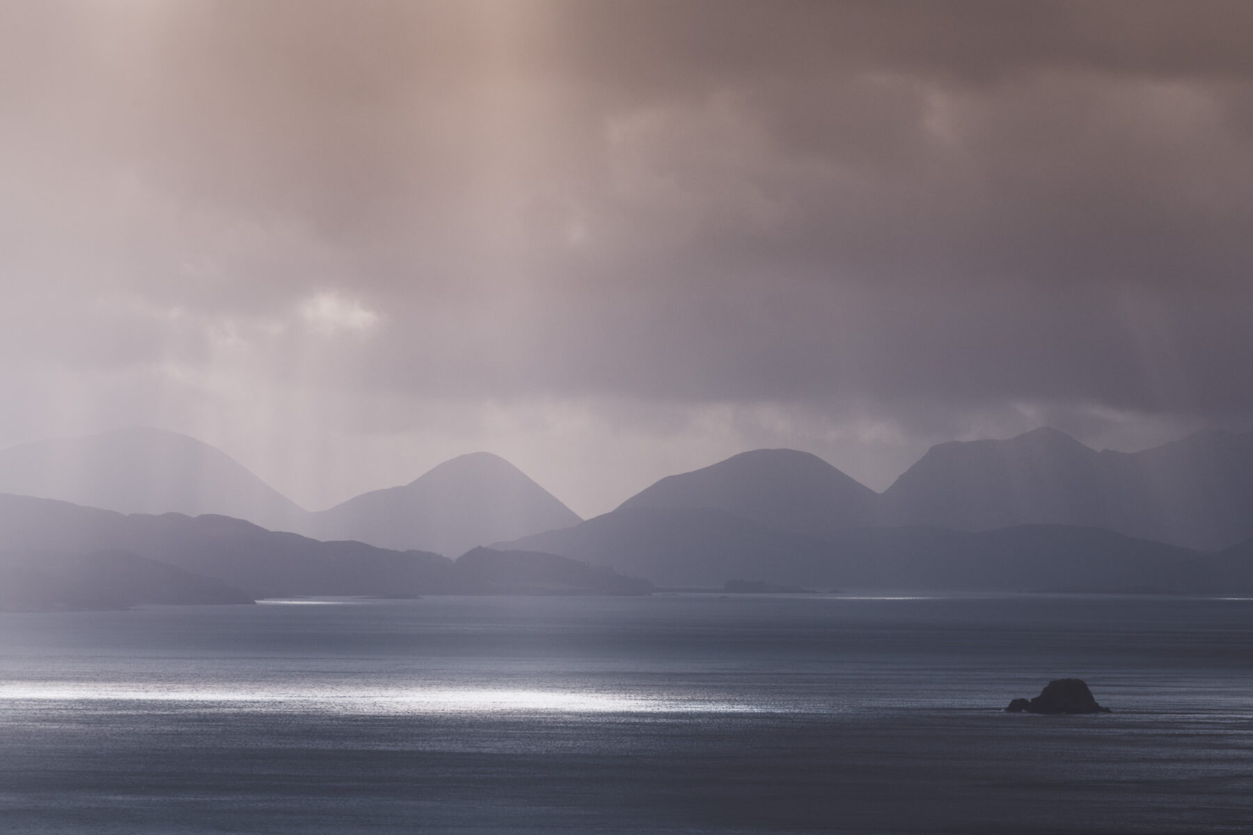 Skye seascape with distant mountains, rain fall and pockets of sunlight on dark dramatic water.