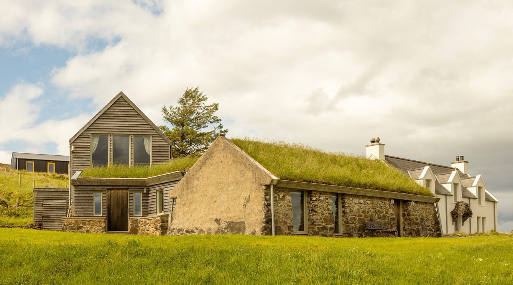 Mint Croft luxury holiday cottages, Isle of Skye. A collection of restored historic buildings with contemporary additions. In front, a turf-roofed hebridean blackhouse and attached timber-clad two storey cottage. Behind, a traditional whitewashed croft house, with three dormer windows upstairs and a heather-covered front porch.