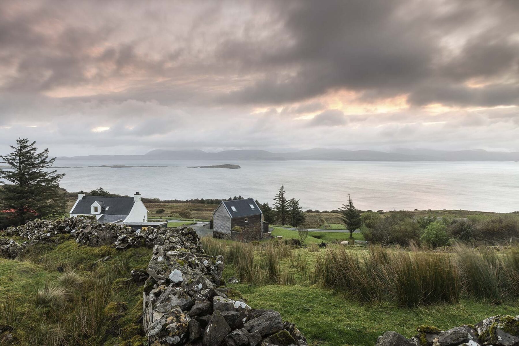 Skye croft, with drystone wall amongst reeds and grass, traditional and contemporary Croft House buildings including a whitewashed stone croft house. Views to the sea.