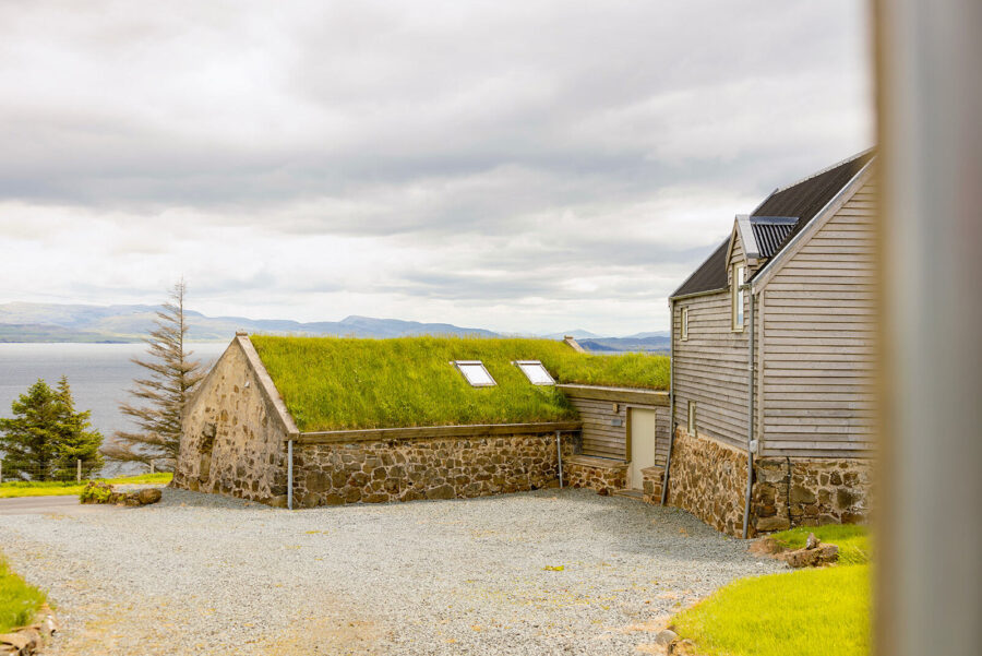 Behind the blackhouse cottage at Mint Croft, Skye. Traditional turf roof on the low stone building with two skylights. The blackhouse is connected to a timber-clad extension with pitched roof. Behind, Loch Snizort and Trotternish.