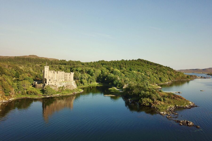 Dunvegan Castle, Skye, seen from the water. Perched above the shore, a historic castle surrounded by woodland on three sides.