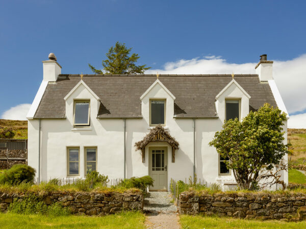 Traditional Scottish Highland croft house cottage at Mint Croft, Skye. A stone whitewashed house with three dormer windows upstairs, a pitched slate roof and a heather-roofed porch above the front door. Set behind a herb garden with low dry stone wall. A bright early summer day with blue sky